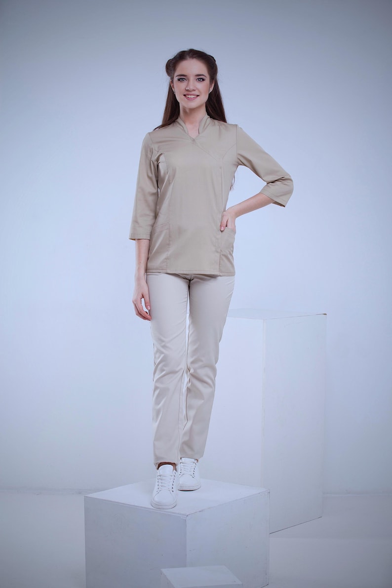 May include: A beige uniform set, including a top with three-quarter sleeves and a V-neck, paired with matching pants. The outfit is worn with white sneakers. The model is standing on a white cube against a light background.