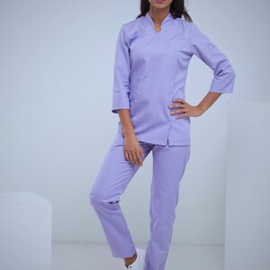 May include: A light purple medical uniform set. The set includes a top with three-quarter sleeves and matching pants. The model is wearing white sneakers. The uniform is made of a smooth, wrinkle-resistant fabric.