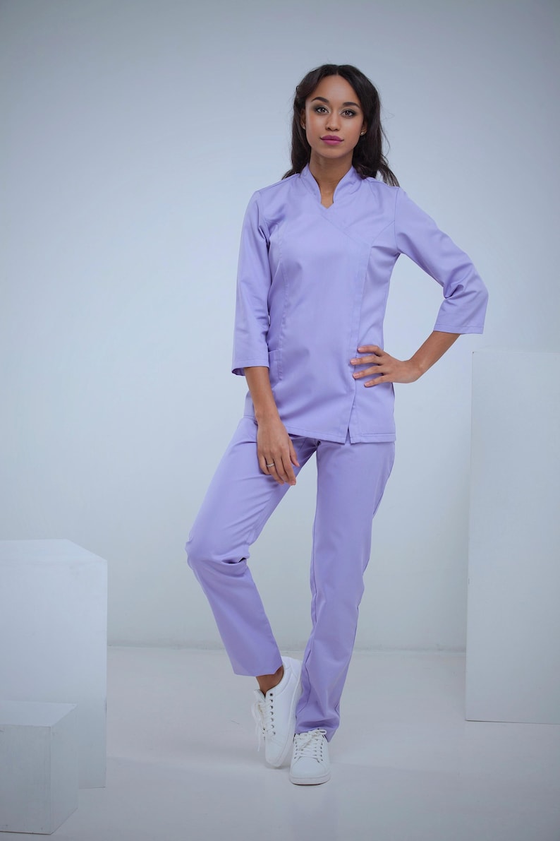 May include: A light purple medical uniform set. The set includes a top with three-quarter sleeves and matching pants. The model is wearing white sneakers. The uniform is made of a smooth, wrinkle-resistant fabric.