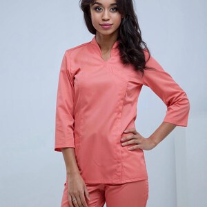 May include: A coral-colored medical uniform set, including a top with a V-neck and three-quarter sleeves, and matching pants. The outfit is made of a smooth fabric and is designed for comfort and functionality in a healthcare setting.
