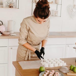 Puede incluir: Una persona con una chaqueta de chef beige y guantes negros est&aacute; decorando merengues blancos sobre papel de hornear en una encimera de madera. Un plato de postres rosas y manzanas verdes tambi&eacute;n est&aacute;n en la encimera.