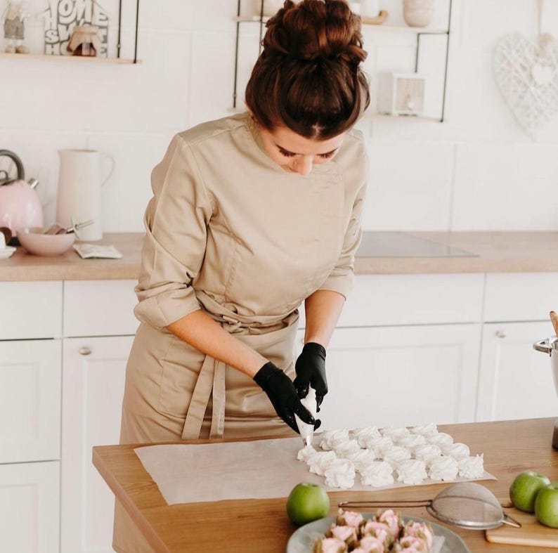 Puede incluir: Una persona con una chaqueta de chef beige y guantes negros est&aacute; decorando merengues blancos sobre papel de hornear en una encimera de madera. Un plato de postres rosas y manzanas verdes tambi&eacute;n est&aacute;n en la encimera.