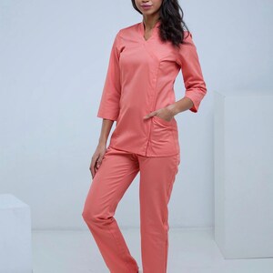 May include: A coral-colored medical uniform set, including a V-neck top with three-quarter sleeves and pockets, paired with matching straight-leg pants. The outfit is made of a smooth fabric and is worn with white sneakers.