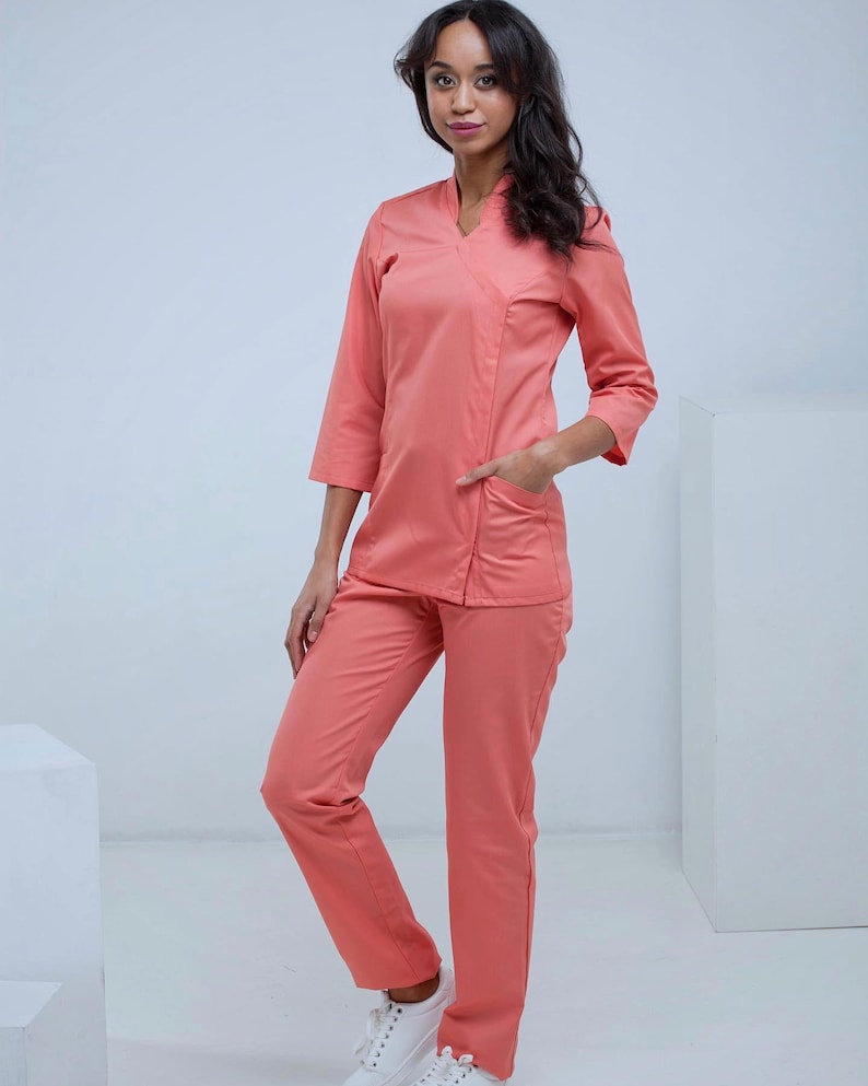 May include: A coral-colored medical uniform set, including a V-neck top with three-quarter sleeves and pockets, paired with matching straight-leg pants. The outfit is made of a smooth fabric and is worn with white sneakers.