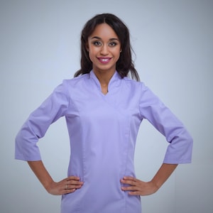 May include: A light purple medical uniform with a wrap-style top and three-quarter sleeves. The uniform has a V-neck and side pockets. The person is smiling and has their hands on their hips.