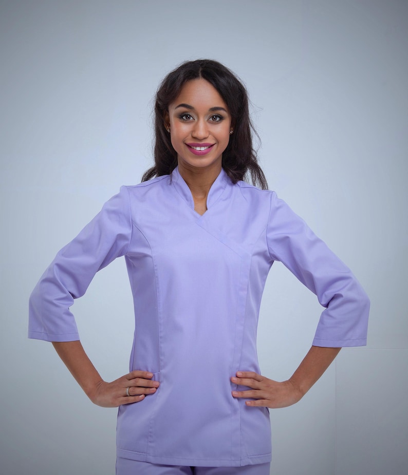 May include: A light purple medical uniform with a wrap-style top and three-quarter sleeves. The uniform has a V-neck and side pockets. The person is smiling and has their hands on their hips.
