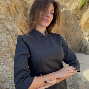 Op de afbeelding: Een zwart topje met een opstaande kraag en driekwartmouwen. Het topje heeft een getailleerd silhouet. De persoon draagt gouden ringen. De achtergrond toont een rotsachtige klif en een zandstrand. Het topje is van glad materiaal.