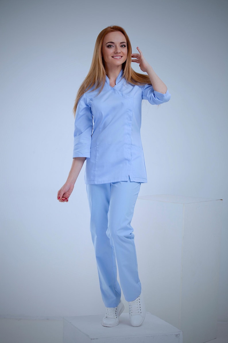 May include: A light blue medical uniform, including a top with a v-neck and three-quarter sleeves, and matching pants. The outfit is worn with white sneakers. The person is smiling and has long, light brown hair.