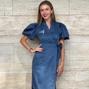 May include: A blue wrap dress with short, puffy sleeves and a tie waist. The dress has a knee-length hemline and a small pocket on the front. The text "IntR" is embroidered on the chest. A decorative pin is attached to the dress.