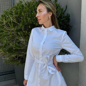 May include: A white shirt dress with long sleeves and a tie waist. The dress has a button-down front and a collared neckline. The dress is knee-length and made of a lightweight fabric. The model is wearing gold earrings.