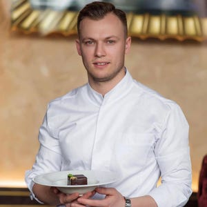 May include: A person in a white chef's coat holds a white plate with a small, square chocolate dessert garnished with a gold leaf. The background is a warm, neutral colour, and the person is smiling at the camera.