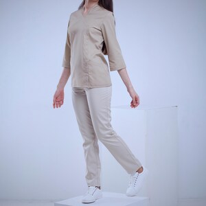 May include: A beige uniform set, including a wrap-style top with three-quarter sleeves and matching pants. The outfit is paired with white sneakers. The model is standing on a white cube against a white backdrop.