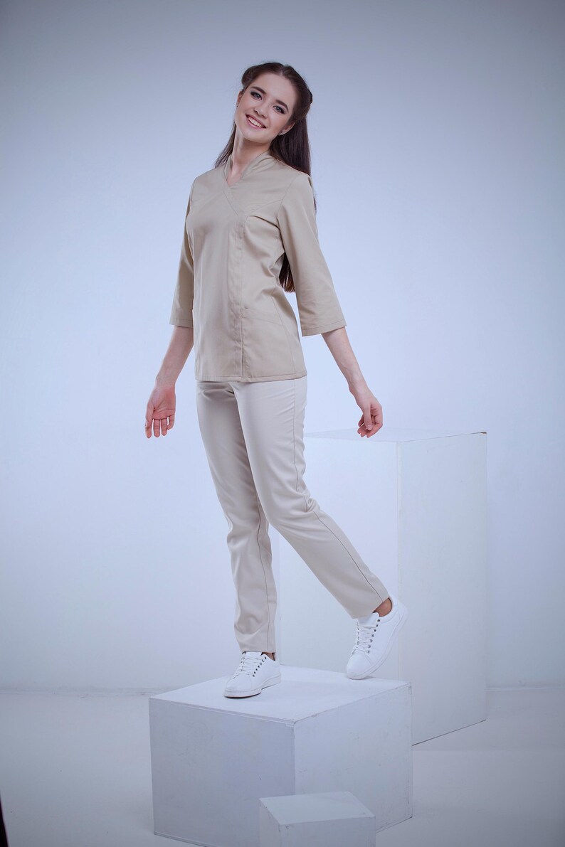 May include: A beige uniform set, including a wrap-style top with three-quarter sleeves and matching pants. The outfit is paired with white sneakers. The model is standing on a white cube against a white backdrop.