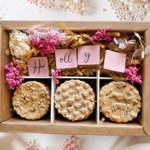 Könnte beinhalten: Eine rechteckige Geschenkschachtel aus Karton mit drei runden, strukturierten Keksen und vier rosa quadratischen Karten, die den Namen Holly und ein Herzsymbol bilden. Die Schachtel ist mit braunem Papierstroh und getrockneten rosa und weißen Blumen gefüllt.