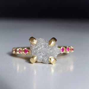 Könnte beinhalten: Ein goldener Ring mit einem großen, grob geschliffenen, grauen Diamanten als Mittelpunkt. Das Band ist mit kleinen, runden, roten Edelsteinen und goldenen Akzenten verziert. Der Ring ist vor einem neutralen Hintergrund platziert.
