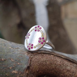 May include: A silver ring featuring a large, oval-shaped face with a brushed matte finish. The surface is decorated with a circular arrangement of small, embedded gemstones, alternating between vibrant pink and pale yellow circular stones.