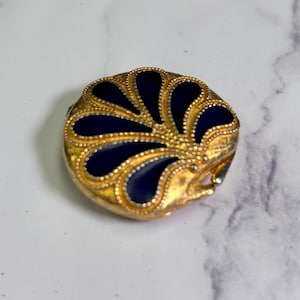 Peut inclure: Un compact rond doré avec un motif floral bleu marine. Le compact a une surface détaillée et texturée et une ouverture à charnière. Les sections bleu marine sont lisses et brillantes, contrastant avec l'or texturé. Un accessoire vintage.