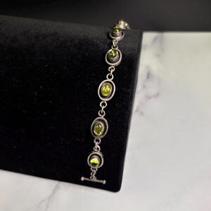 May include: A silver bracelet with oval-shaped green gemstones. The bracelet features a series of linked ovals, each containing a vibrant green stone. The clasp is a simple T-bar design. The bracelet is displayed against a black background.