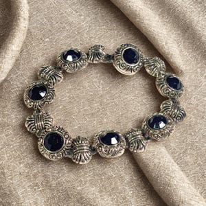 Può includere: Un bracciale color argento caratterizzato da una serie di maglie circolari con intricati motivi a volute. Ogni maglia è incastonata con una pietra cabochon rotonda, liscia e di colore blu marino scuro. Il bracciale alterna segmenti decorativi in metallo.