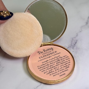 Può includere: Cofanetto dorato aperto con specchio e piumino. Il cofanetto contiene cipria rosa. Il testo sulla cipria recita "Du Barry Cloudsilk Compressed Powder". Un piumino beige è accanto.
