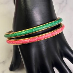 May include: Two bangle bracelets. One is green, the other is pink. Both have a gold-colored metal band and a textured, snake-like pattern. The bracelets are displayed on a black hand-shaped jewelry holder.