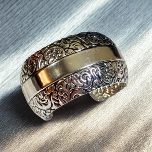 May include: A wide cuff bracelet featuring a polished, smooth gold-tone center band flanked by two textured silver-tone sections. The silver portions display intricate, raised swirling patterns that contrast with the plain, reflective gold-tone stripe.