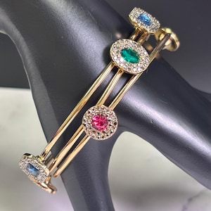 May include: Gold-tone bangle bracelet with three parallel bars, each adorned with a circular or square-shaped jewel. The jewels are in shades of blue, green, and pink, surrounded by smaller clear stones. The bracelet is displayed on a black mannequin hand.