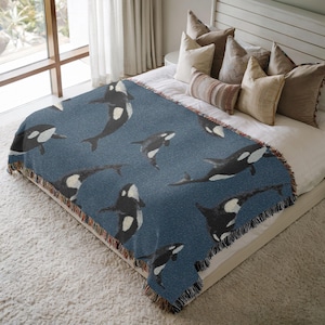 May include: A blue throw blanket featuring a pattern of black and white orca whales. The blanket is fringed on the edges and is displayed on a bed with neutral-colored pillows and a white bedspread. The orcas are in various positions.