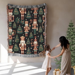 May include: A woven Christmas-themed throw blanket with a black background, featuring nutcrackers and Christmas trees in shades of brown, green, and red. The blanket has a fringed edge and is displayed on a wall.