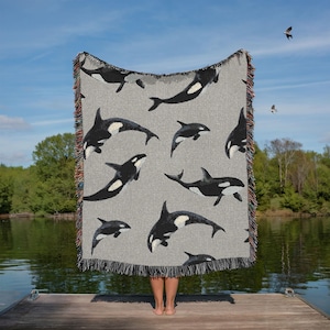 May include: A gray woven throw blanket featuring a pattern of black and white orcas. The blanket has a fringed edge with multiple colors. The blanket is held up by a person standing on a wooden dock.