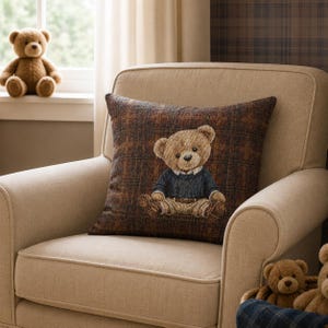 Puede incluir: Un sillón beige con un cojín decorativo con un diseño de oso de peluche. El cojín tiene un fondo a cuadros marrón. Un oso de peluche está sentado en el alféizar de la ventana. Una cesta con osos de peluche está en la esquina inferior derecha.