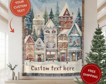 May include: Woven throw blanket featuring a winter village scene with snow-covered buildings and evergreen trees. The blanket has a fringed edge and the text "Custom text here" is displayed. The color palette includes blues, browns, and whites.