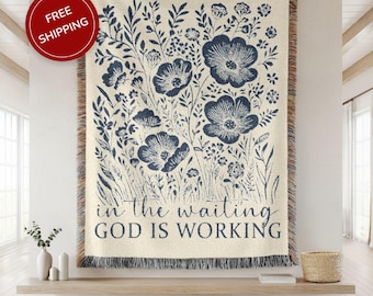May include: Cream-coloured woven throw blanket with a navy blue floral design and the text "in the waiting GOD IS WORKING." The blanket has a fringed edge and is displayed on a wooden shelf.
