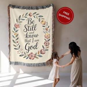 May include: Cream-coloured woven throw blanket with the text "Be Still and know that I am God" surrounded by a floral wreath. The blanket has fringed edges and is hanging on a wall.