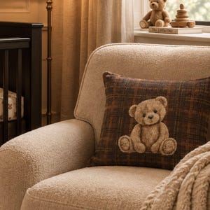 Puede incluir: Un cojín cuadrado con un patrón de cuadros en marrón oscuro y negro, que presenta una aplicación texturizada centrada de un oso de peluche marrón claro sentado. El cojín está colocado sobre un sillón texturizado de color crema.