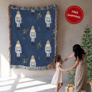 May include: A blue woven throw blanket with a repeating pattern of light blue nutcracker figures and green holly leaves. The blanket has a fringed edge and is displayed on a wall.