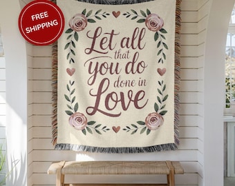 May include: Beige woven throw blanket featuring the text "Let all that you do done in Love" framed by floral and heart motifs. The blanket has a fringed edge and hangs on a white wall above a wooden bench.