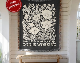 May include: Black woven throw blanket with a cream floral design and the text "in the waiting GOD IS WORKING." The blanket has a fringed edge and is hanging on a white wall above a wooden bench.