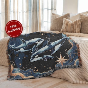 May include: A woven throw blanket featuring a dark blue background with a design of orca whales, stars, and celestial elements. The blanket has a fringed edge and is draped over a bed with neutral-toned pillows.