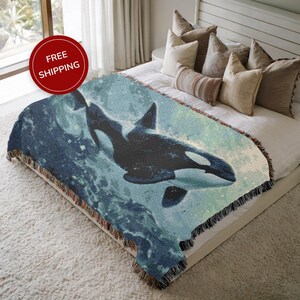 May include: A woven throw blanket featuring a black and white orca whale design against a blue and teal background. The blanket has a fringed edge and is displayed on a bed with pillows.