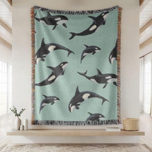 May include: A woven throw blanket featuring a repeating pattern of black and white orcas on a light green background. The blanket has a fringed edge and is displayed on a wall. The orcas are in various poses, creating a dynamic design.