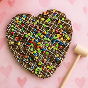 May include: A heart-shaped chocolate dessert with white icing lattice and colorful candy toppings. A small wooden mallet rests on a pink surface with heart patterns, suggesting a treat designed to be broken.
