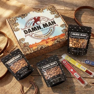 May include: A cowboy-themed snack starter kit with a box and several bags of nuts and meat sticks. The box features the text "DAMN MAN" and "Cowboy Starter Kit". The bags are labeled "Texas Peanuts", "Memphis Almonds", and "Whiskey Nut Mix".