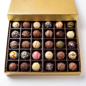 May include: A gold box filled with an assortment of chocolate truffles. The truffles are round and come in various colors and decorations, including sprinkles, stripes, and chocolate shavings. The box is open, revealing the arrangement of treats.
