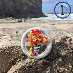 May include: An embroidered brooch with a floral design. The brooch features a bouquet of red, orange, and yellow flowers within a white circular frame. The background shows a beach and ocean. The brooch is set on a rocky surface.