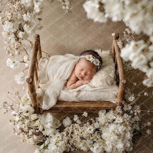 May include: A newborn baby sleeps peacefully in a small wooden bed, draped with a soft white blanket and pillow. The bed is surrounded by a wreath of white flowers and greenery, creating a serene and natural setting. The baby wears a floral headband.