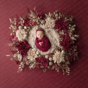 May include: A newborn baby wrapped in a deep red blanket and headband, nestled in a cream-colored nest. The baby is surrounded by a wreath of burgundy, cream, and gold flowers and ornaments, set against a deep red background.