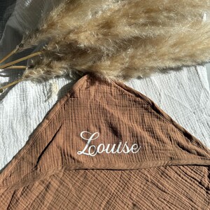 Puede incluir: Una toalla con capucha marr&oacute;n para beb&eacute; con el nombre "Louise" bordado en escritura blanca. La toalla est&aacute; hecha de una tela texturizada y est&aacute; doblada. Hierba de la pampa seca en el fondo.