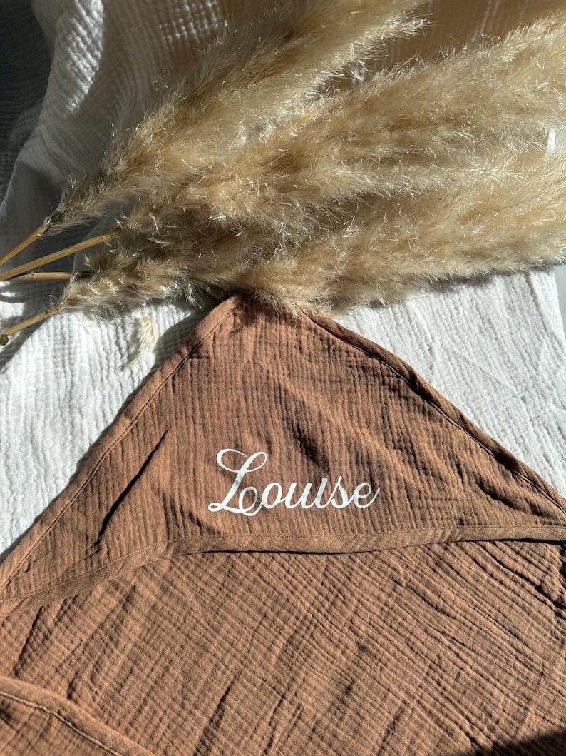 Puede incluir: Una toalla con capucha marr&oacute;n para beb&eacute; con el nombre "Louise" bordado en escritura blanca. La toalla est&aacute; hecha de una tela texturizada y est&aacute; doblada. Hierba de la pampa seca en el fondo.