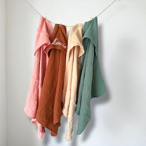 Puede incluir: Varias toallas con capucha para beb&eacute;s en tonos rosa, marr&oacute;n, beige y verde, colgadas de una cuerda. La toalla marr&oacute;n tiene bordado el nombre "Louise" en blanco. Las toallas parecen estar hechas de un material suave y texturizado.